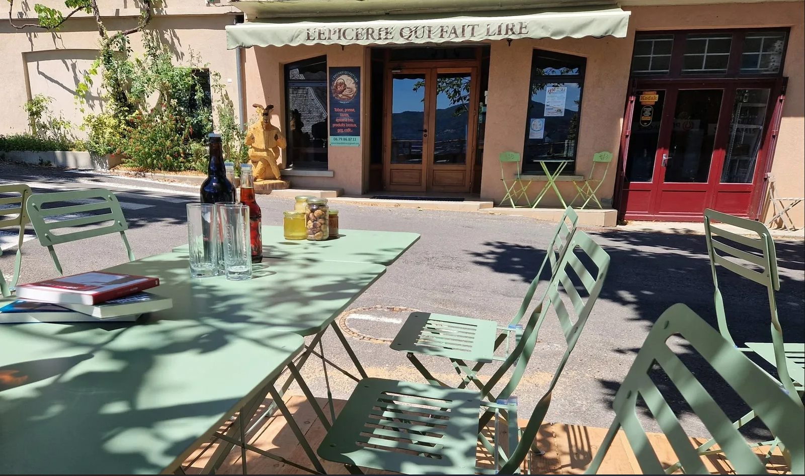 Vue depuis la terrasse de L'Épicerie qui fait lire : une table verte avec des livres, des bouteilles et des pots de miel, et en face le magasin avec son store « L'Épicerie qui fait lire » et la chèvre en bois devant la vitrine